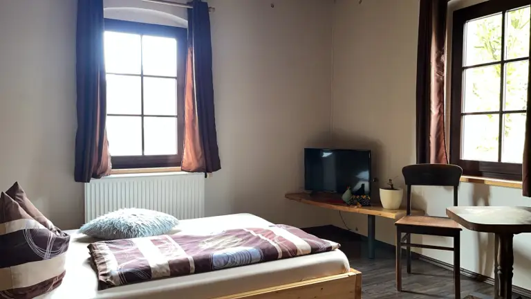 Helles Doppelzimmer der Pension Deftige Pfanne mit Holzbett, zwei Fenstern mit dunklen Vorhängen und kleinem Tisch mit Stuhl