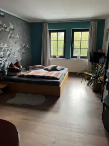 Schlafzimmer der Ferienwohnung: großes Holzbett vor einer Tapete mit weißen Vögeln und Zweigen, zwei Fenster mit Blick ins Grüne