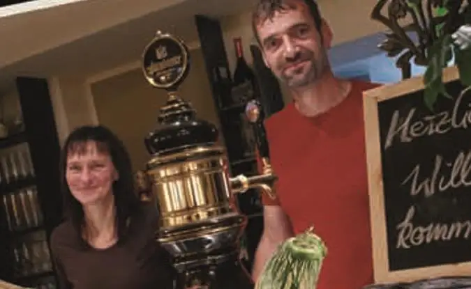 Die Gastgeber des Gasthofs Deftige Pfanne lachend hinter dem Tresen der Gaststube, neben ihnen eine Kreidetafel mit einem Willkommensgruß