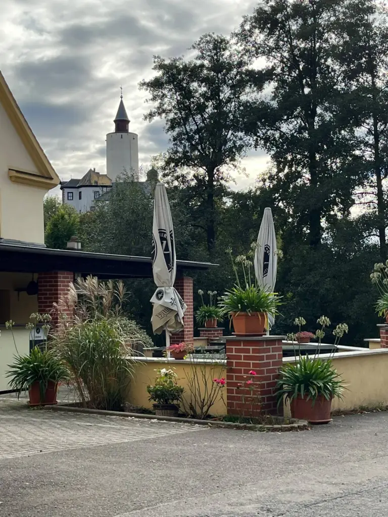 Sonnenterrasse des Gasthofs Deftige Pfanne mit Blumenkübeln und Sonnenschirmen, dahinter ragt der Turm der Burg Posterstein über die Bäume